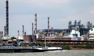 Refinería industrial con múltiples chimeneas y estructuras a lo largo de un canal de agua, con una barcaza pasando cerca.