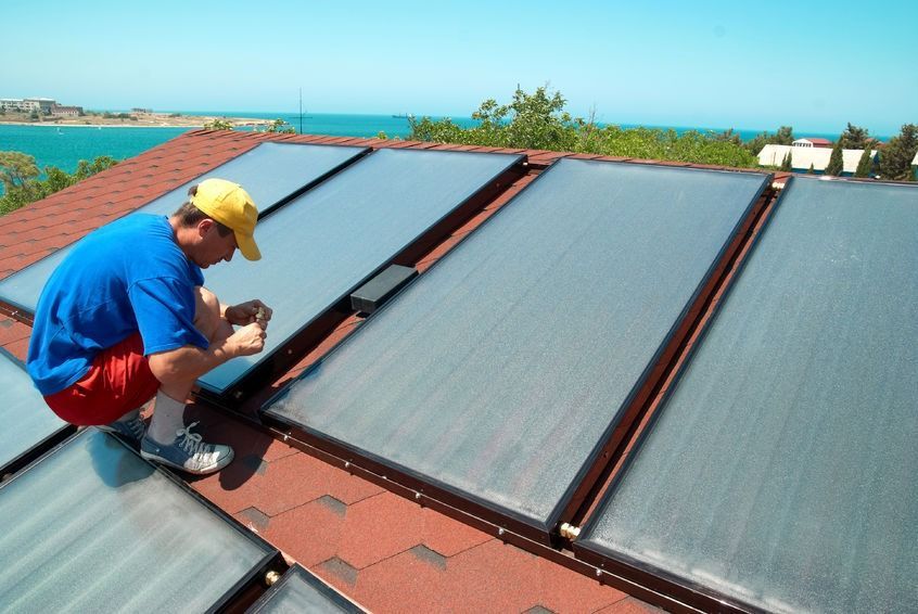 Un hombre con una camisa azul y un sombrero amarillo está trabajando en paneles solares en un techo.