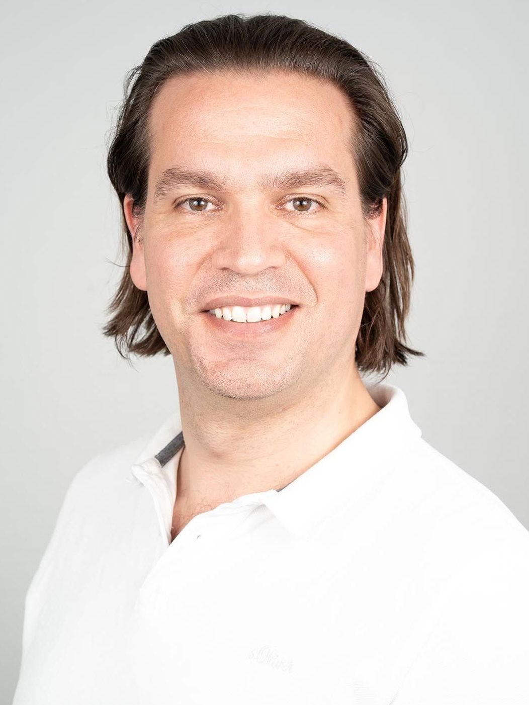 Ein Mann mit langen Haaren trägt ein weißes Hemd und lächelt in die Kamera.