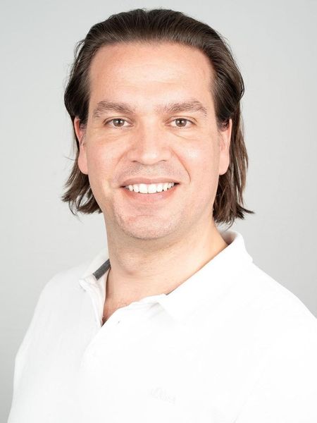 Ein Mann mit langen Haaren trägt ein weißes Hemd und lächelt in die Kamera.