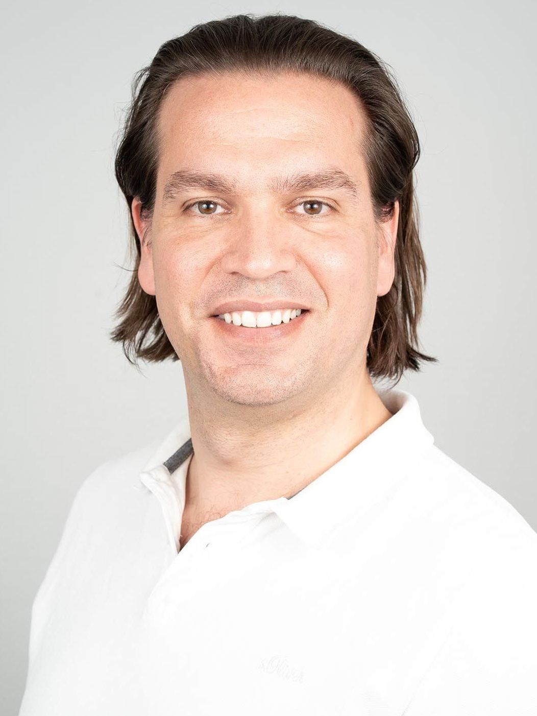 Ein Mann mit langen Haaren trägt ein weißes Hemd und lächelt in die Kamera.