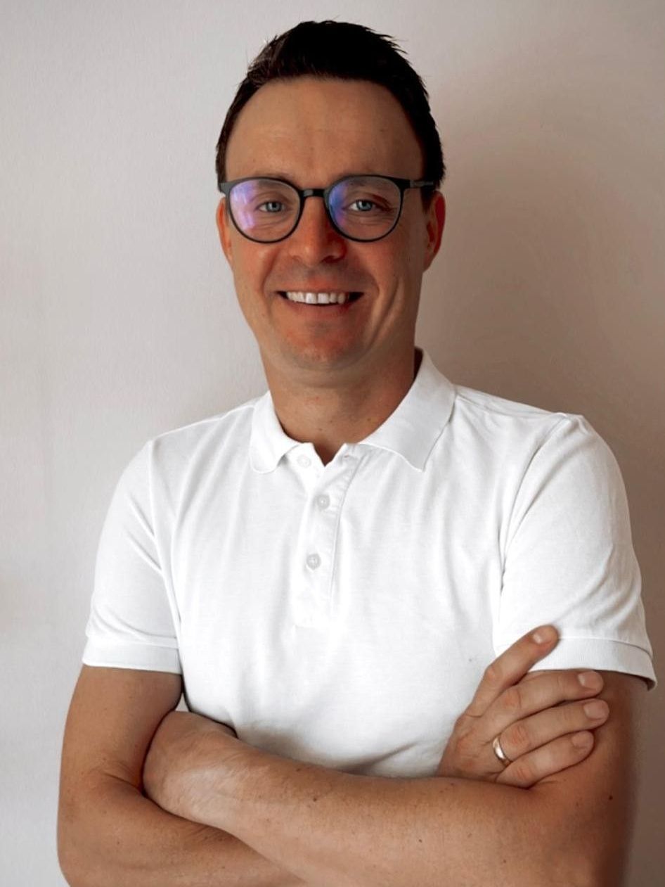 Ein Mann mit Brille und weißem Hemd lächelt und verschränkt die Arme.