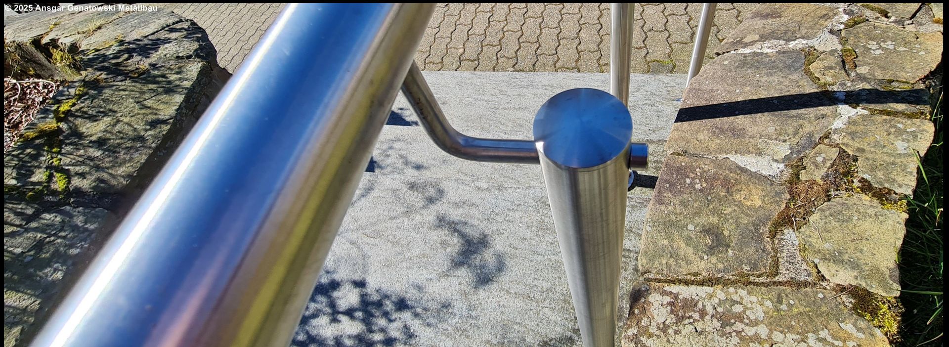 Metallgeländer mit abgerundeter Oberseite und gebogener Stütze über einer Steinoberfläche.