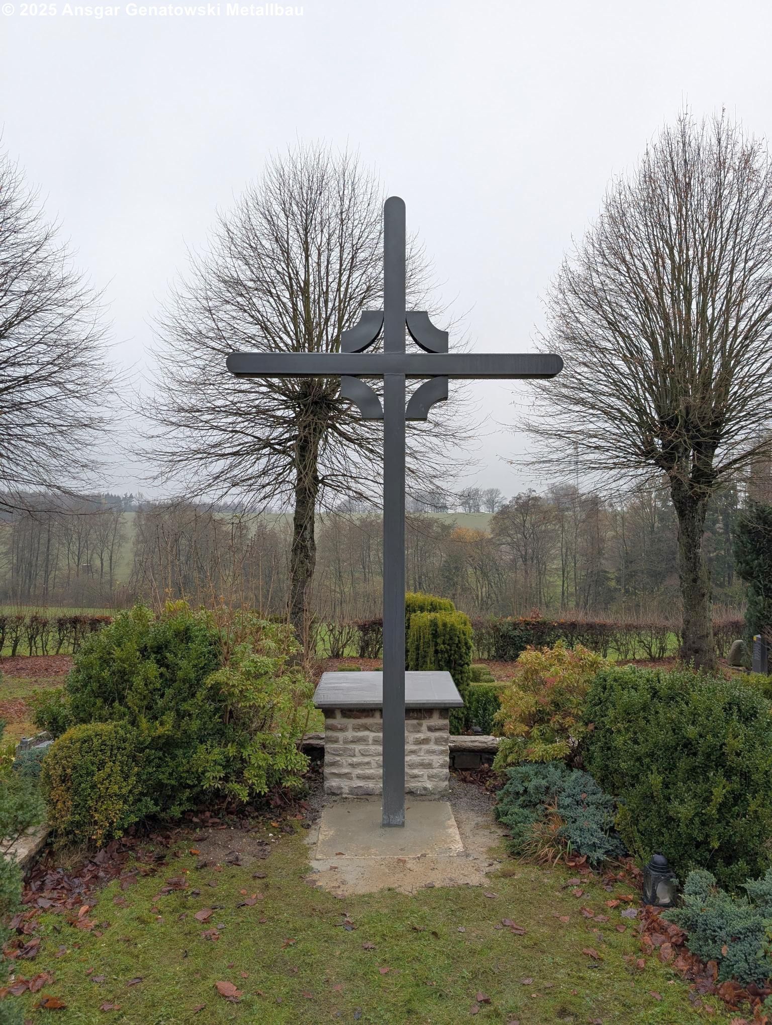 Graues Kreuz als Grabstein auf einem Backsteinsockel, umgeben von Büschen und Bäumen, unter einem bedeckten Himmel.