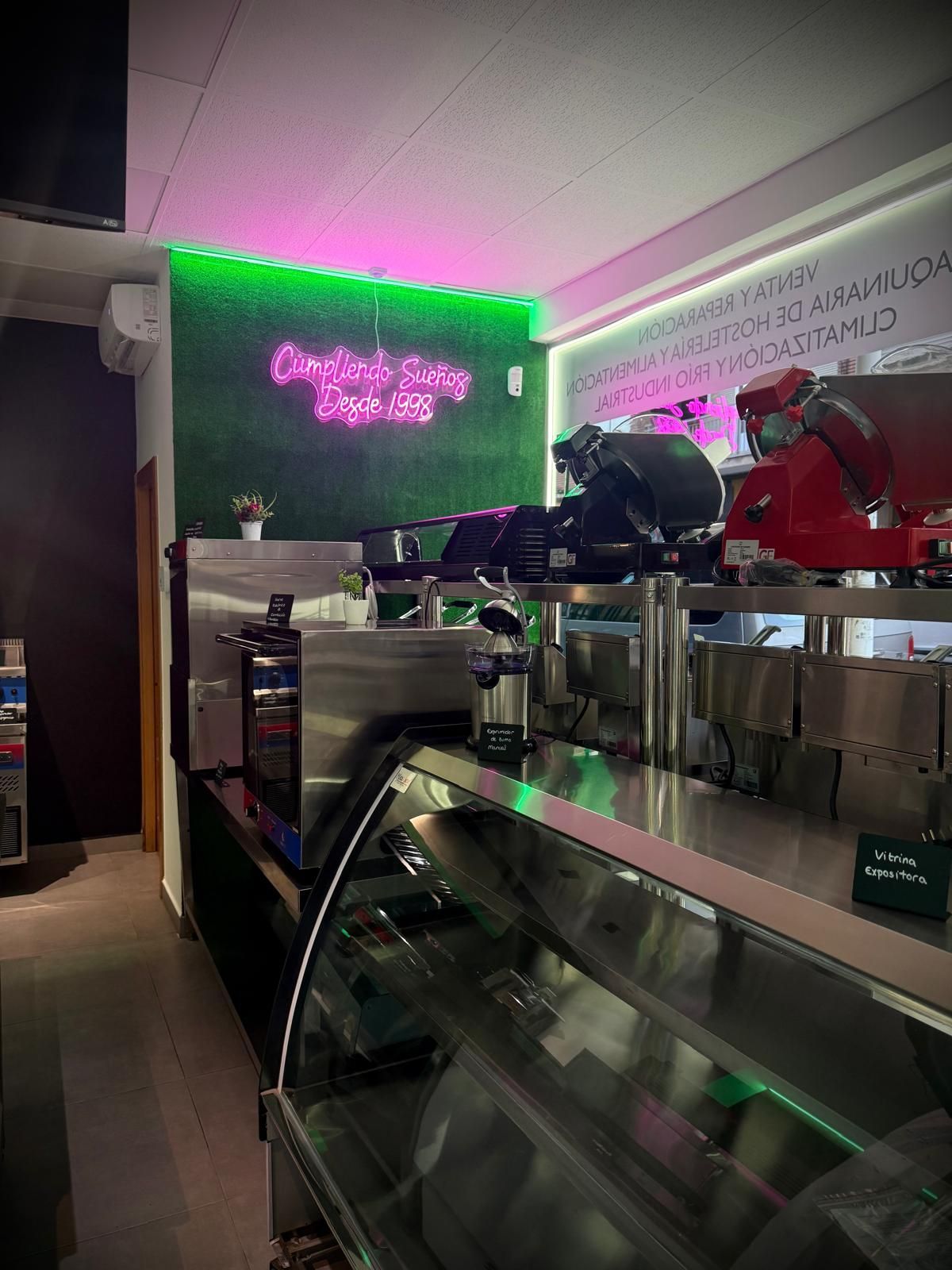 Interior de cafetería moderno con vitrina de cristal, máquinas de café e iluminación de neón rosa verdoso.