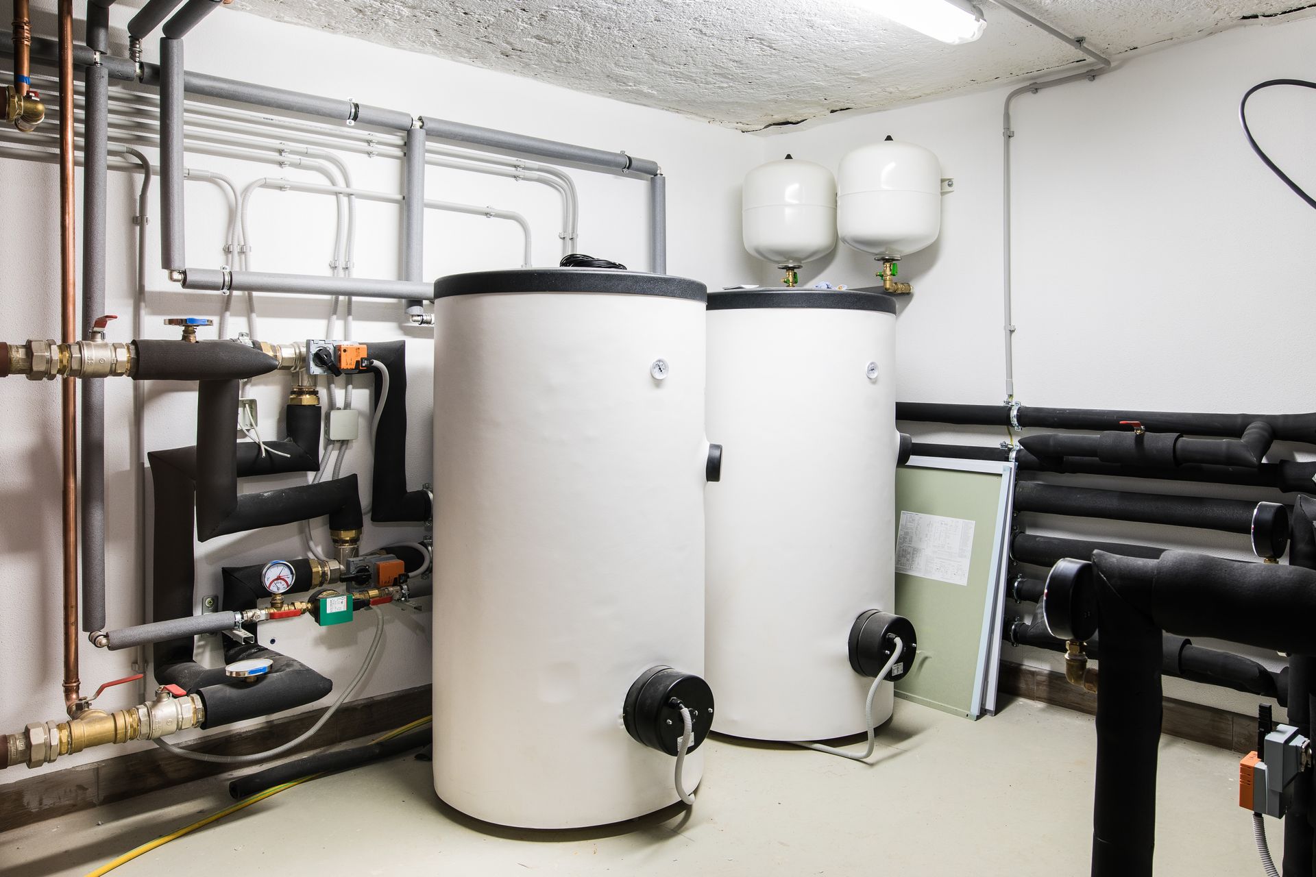 Local technique au sous-sol avec chauffe-eau blancs, tuyaux et conduites de chauffage isolées noires.