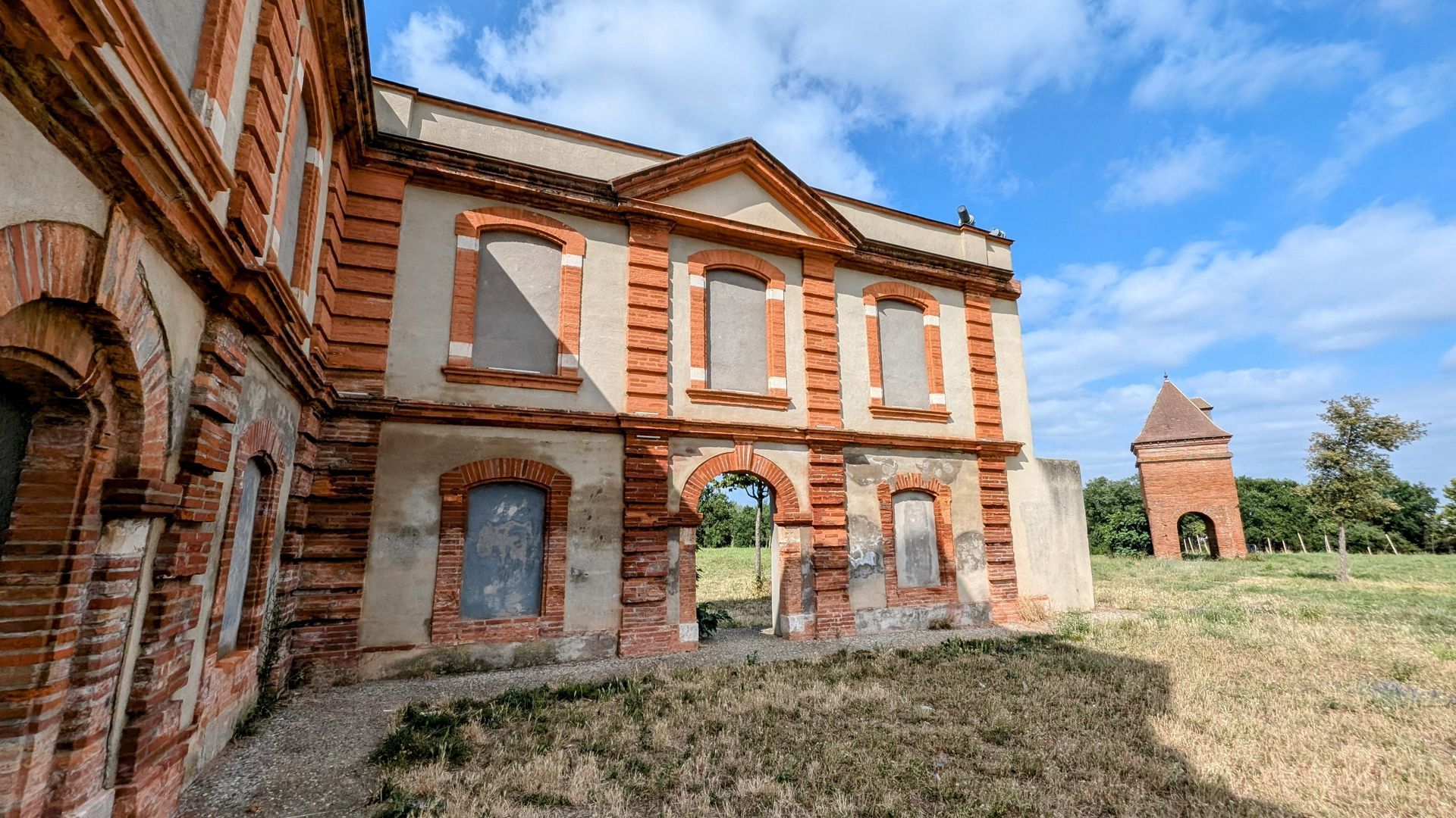 Bâtiment abandonné en briques rouges, avec des fenêtres cintrées et une petite tour en briques, dans un champ herbeux sous un ciel bleu.