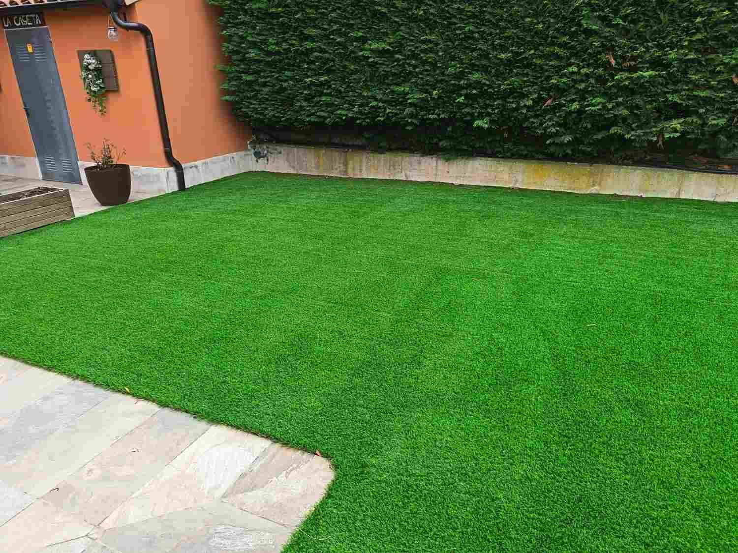 Césped verde junto a un edificio naranja y un seto alto, con un patio de baldosas en primer plano.