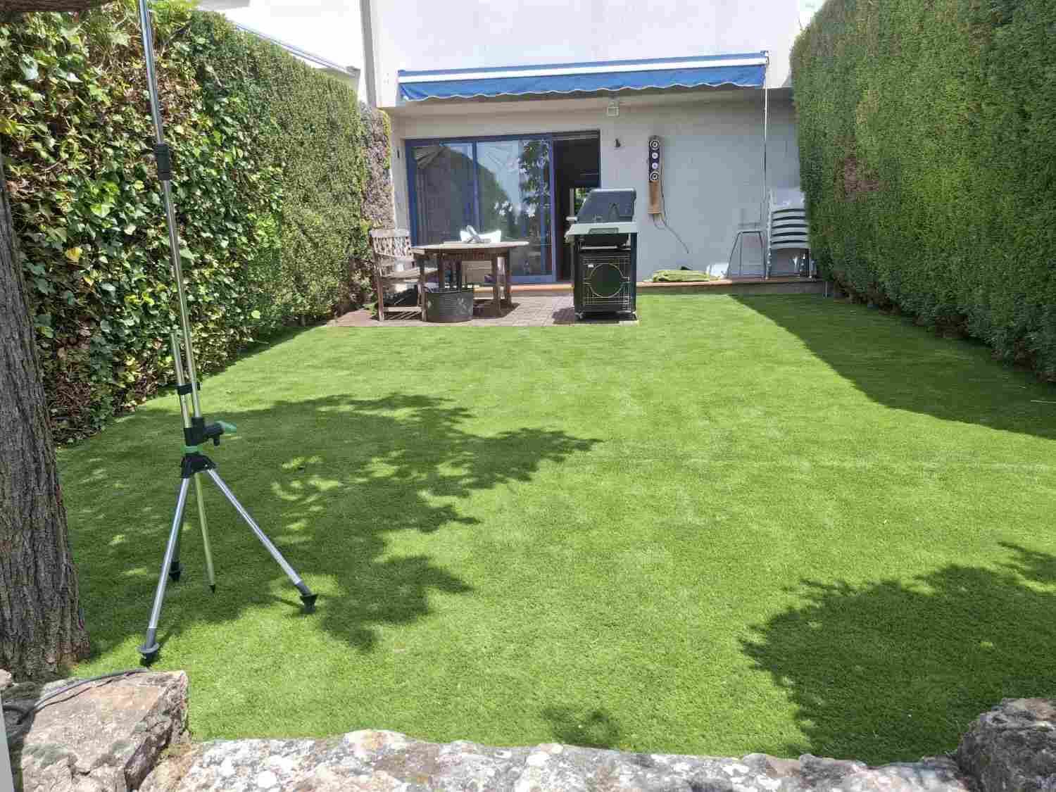 Patio trasero con césped artificial, setos, mesa de patio, parrilla para barbacoa y un trípode cerca del césped.