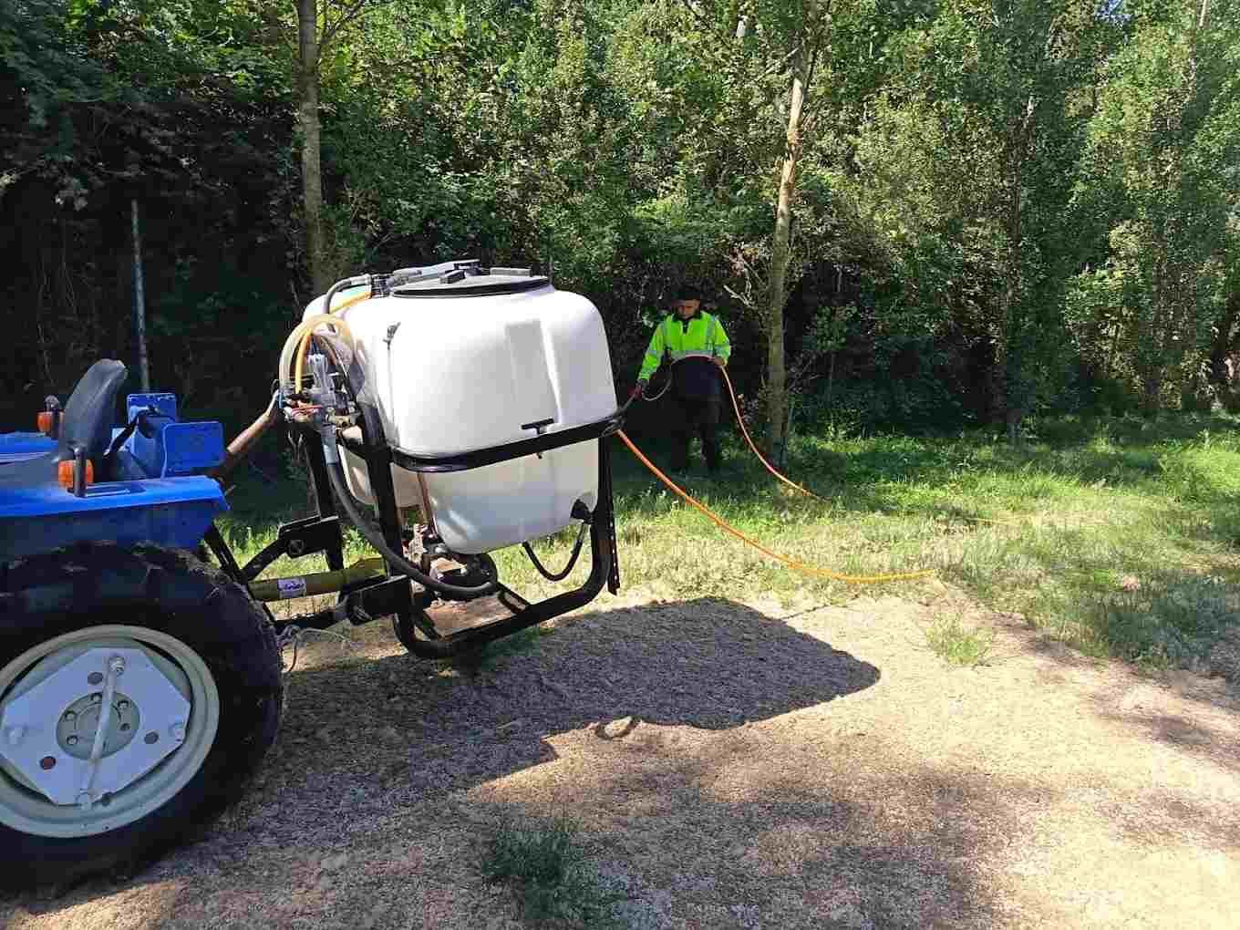Un tractor azul rocía agua en un sendero boscoso; un trabajador con un chaleco reflectante.