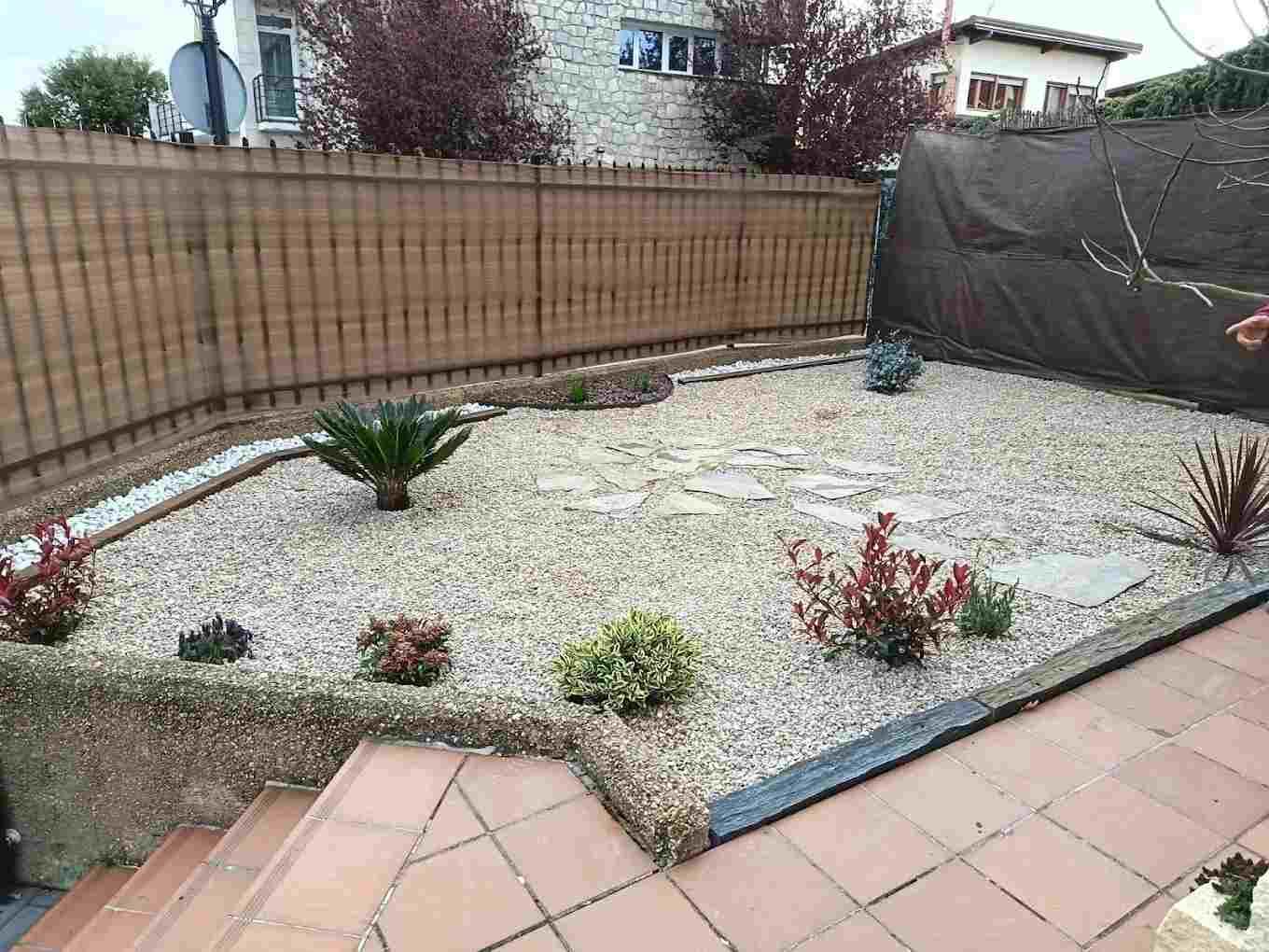 Pequeño jardín de grava con plantas suculentas, valla de madera y baldosas de terracota en el patio.