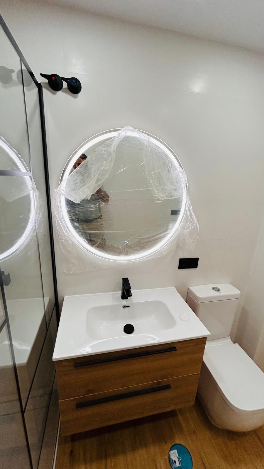 Baño moderno con tocador de madera, espejo redondo retroiluminado, grifería negra e inodoro blanco.