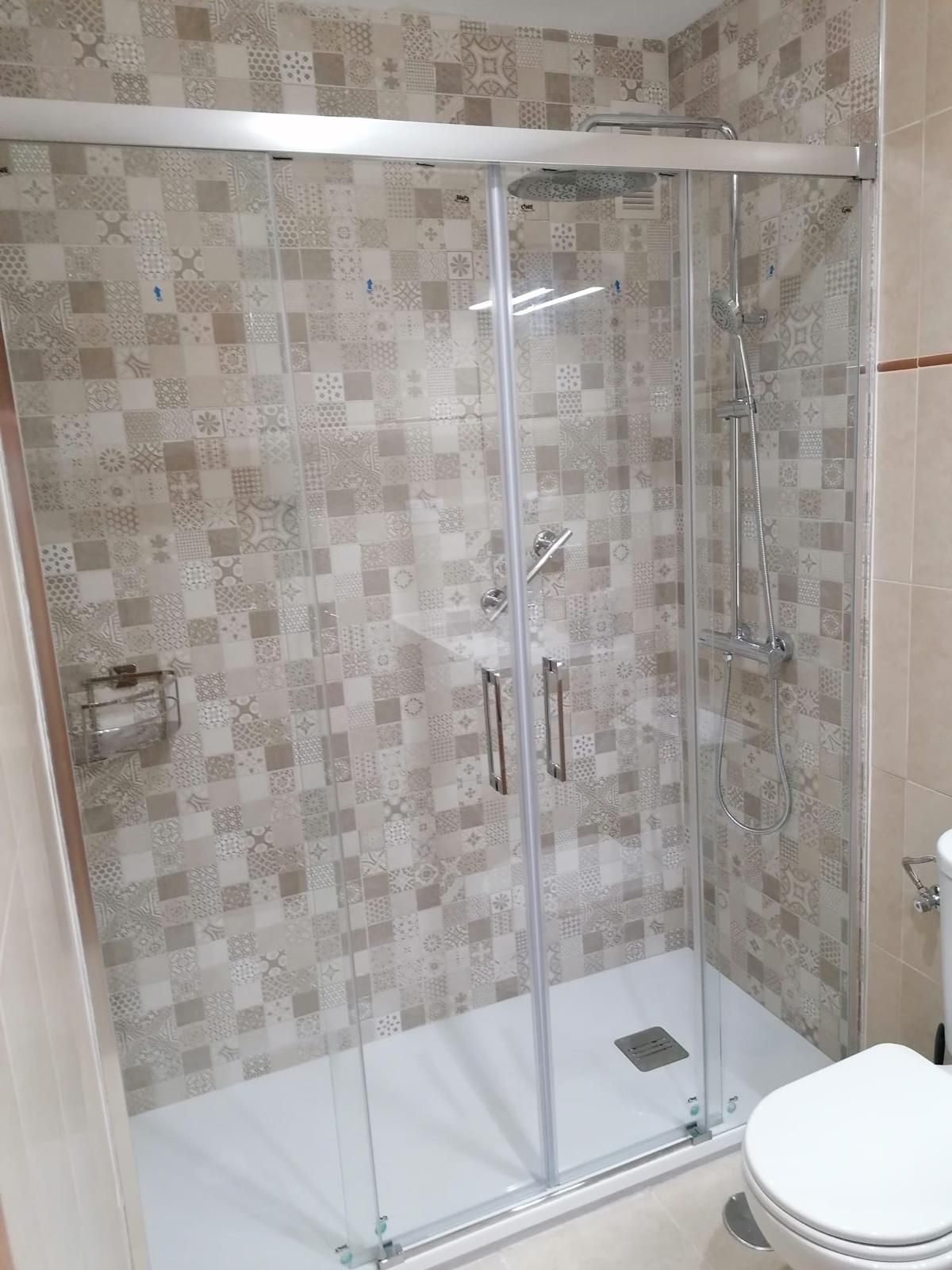 Cabina de ducha con puertas de cristal, paredes alicatadas y base blanca, junto a un inodoro.