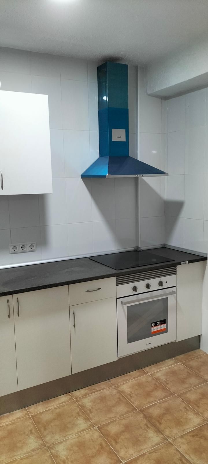 Cocina con gabinetes claros, campana extractora azul, encimera negra, horno y piso de baldosas.