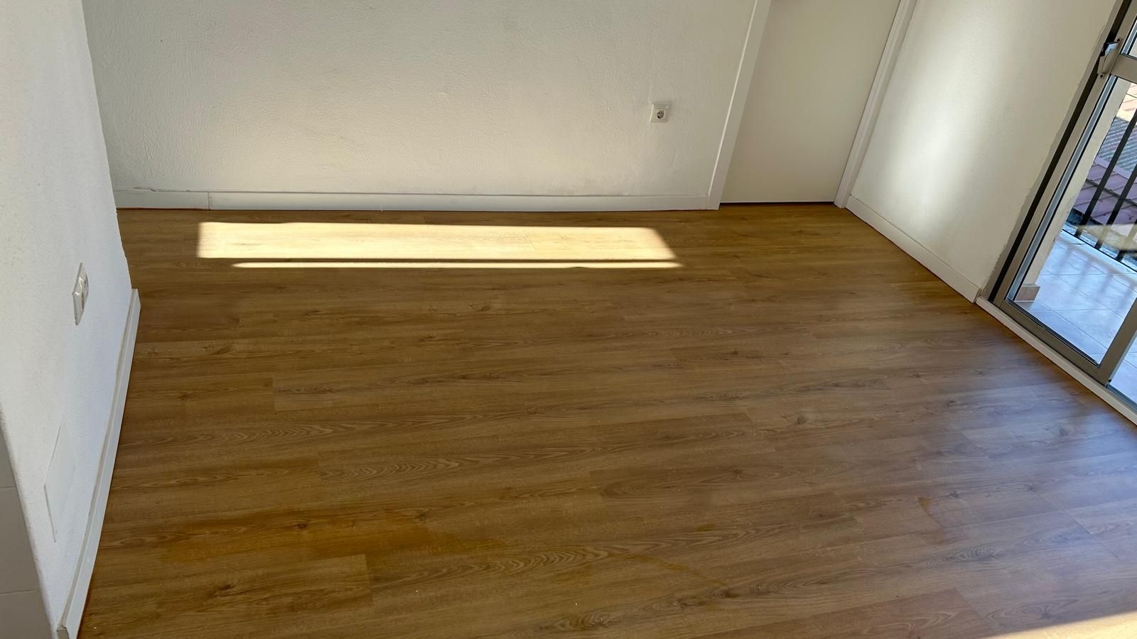 Habitación vacía con suelo imitación madera y paredes blancas. La luz del sol se filtra por el suelo.