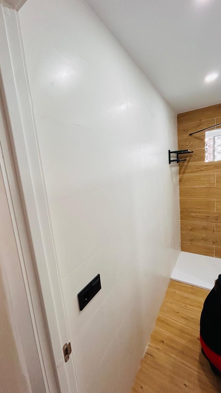 Pared blanca con enchufe y toallero negros. Suelo de madera y pared de azulejos marrones visibles al fondo.