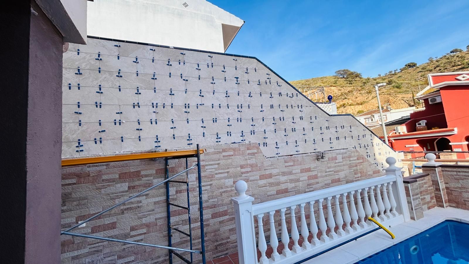 Muro exterior con aislamiento y mampostería, andamios, barandilla blanca, cielo azul.
