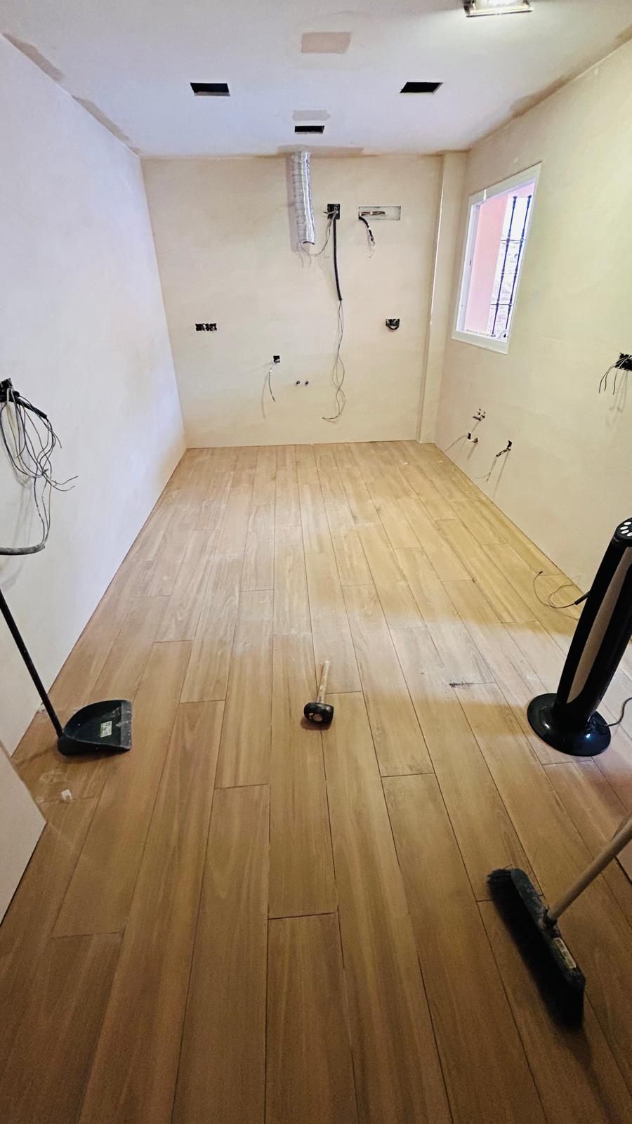 Habitación vacía con suelo de madera clara, paredes sin terminar y cableado eléctrico. Se ven una ventana y herramientas.