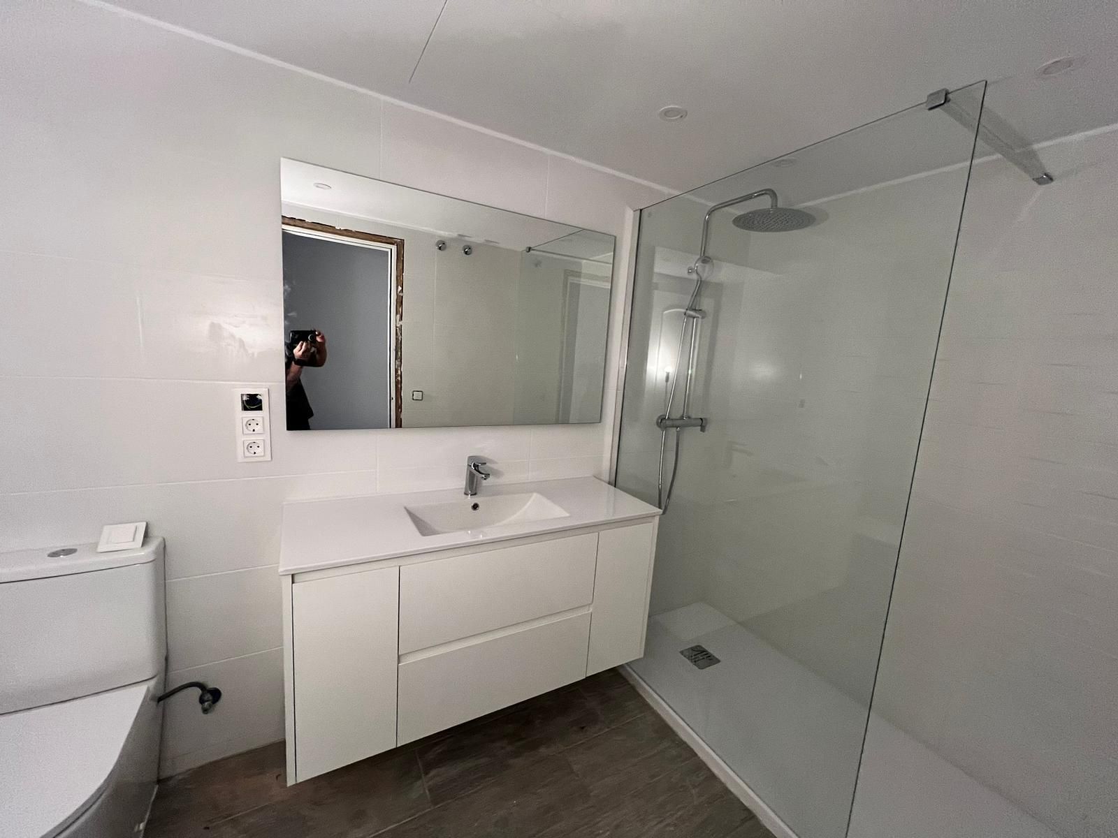 Baño moderno blanco con inodoro, lavabo, espejo y ducha con mampara de cristal.