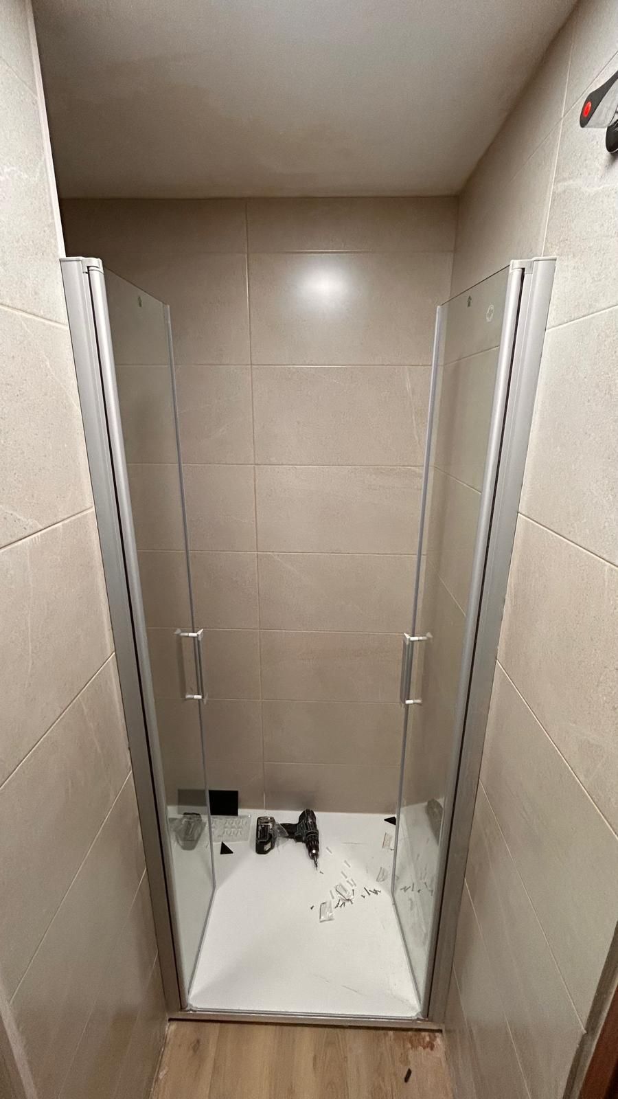 Cabina de ducha con puertas de cristal y base blanca, rodeada de azulejos beige.