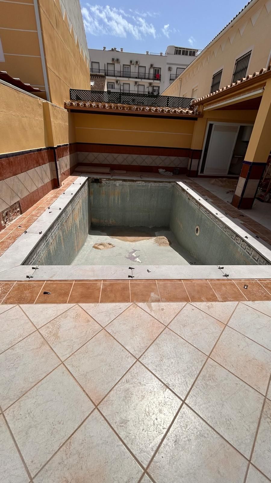 Piscina rectangular vacía rodeada de azulejos estampados y edificios.