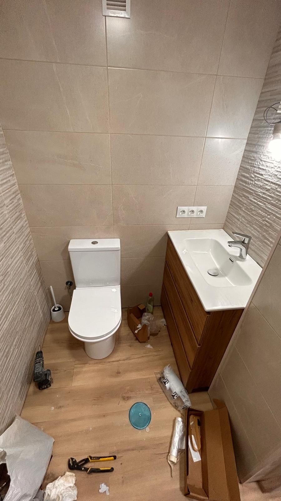 Baño en construcción con inodoro, lavabo y mueble de madera. Paredes de azulejos beige y suelo de madera clara.