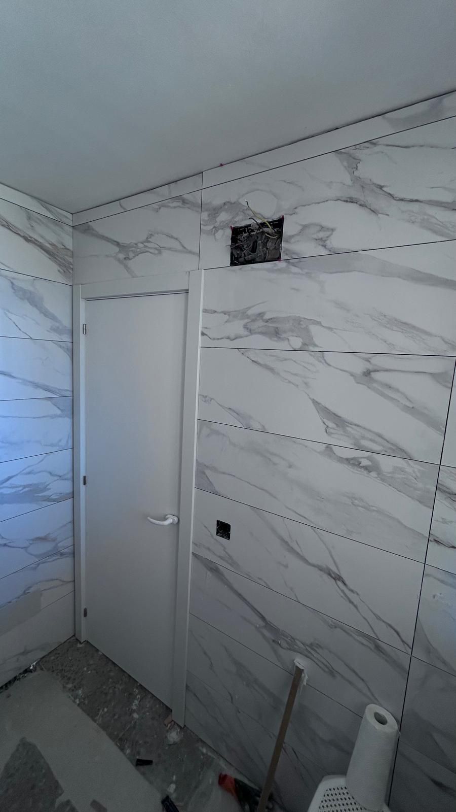 Baño en construcción con azulejo con diseño de mármol, puerta blanca y caja eléctrica expuesta.