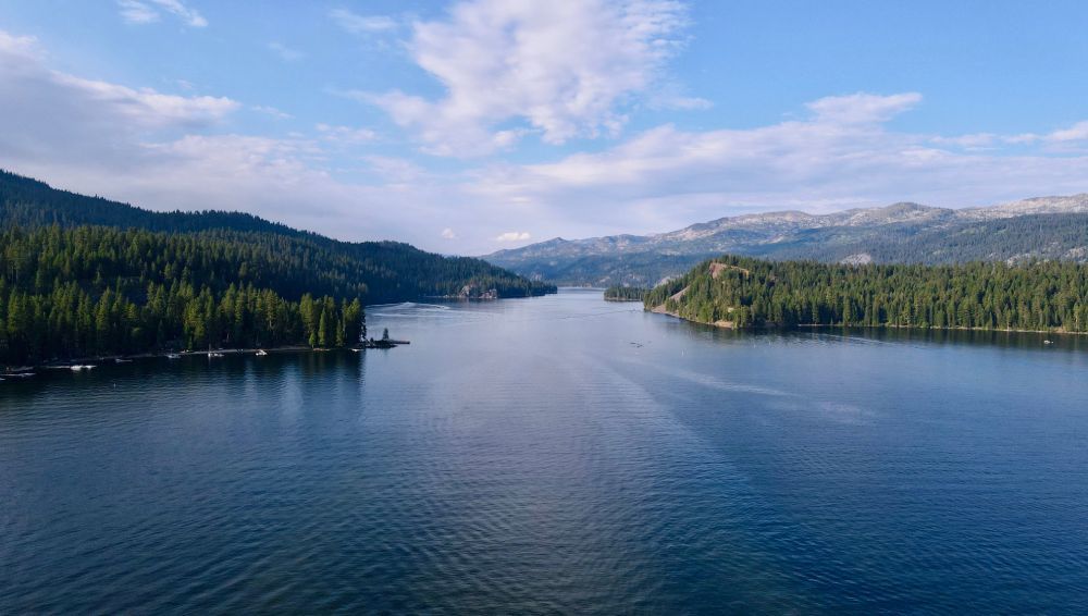 A wide lake surrounded by evergreen forests, under a blue sky with scattered clouds.