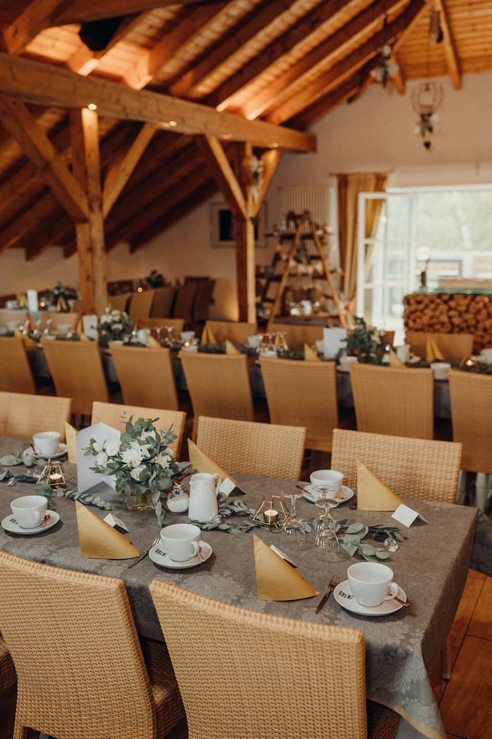Lange Tische sind für ein geselliges Beisammensein in einem rustikalen Interieur mit Holzbalken und einem Fenster aufgestellt.