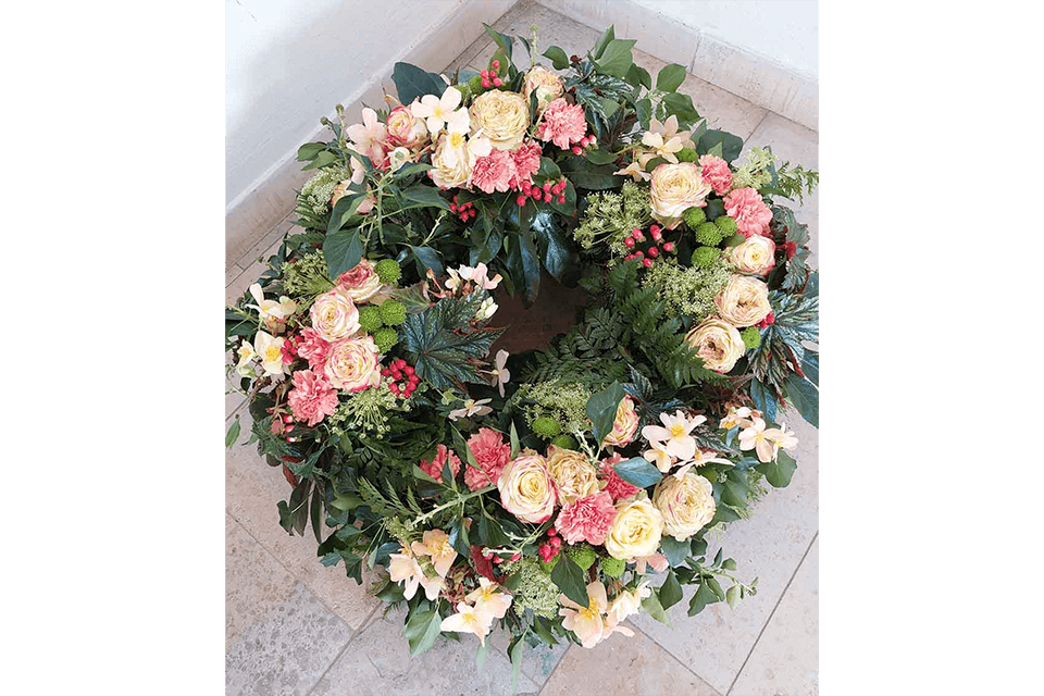 Ein Kranz aus hellrosa, pfirsichfarbenen und weißen Blüten mit grünem Laub auf einem gefliesten Boden.