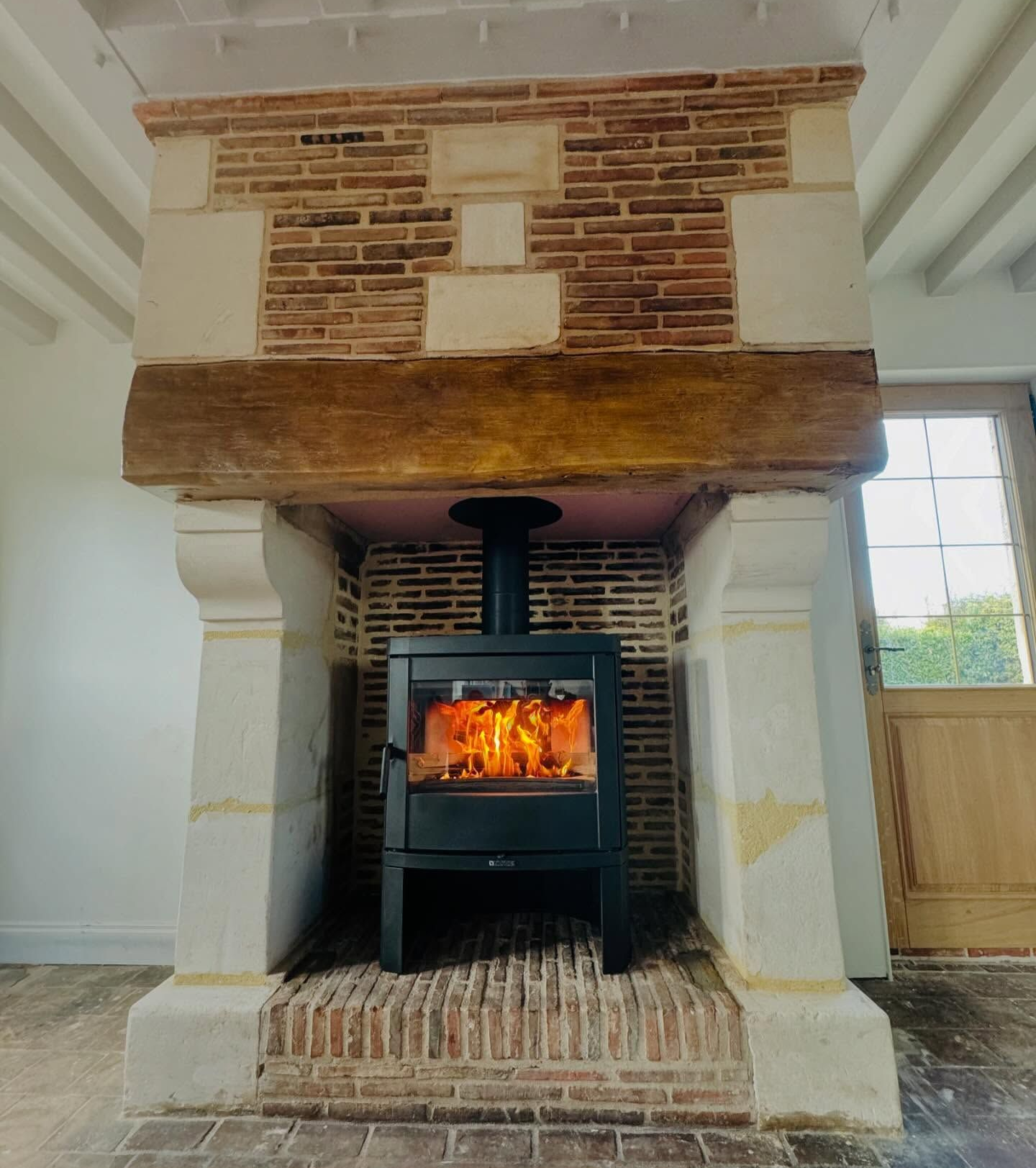 Poêle à bois dans une cheminée en briques et en pierres avec une poutre en bois. Flammes visibles.