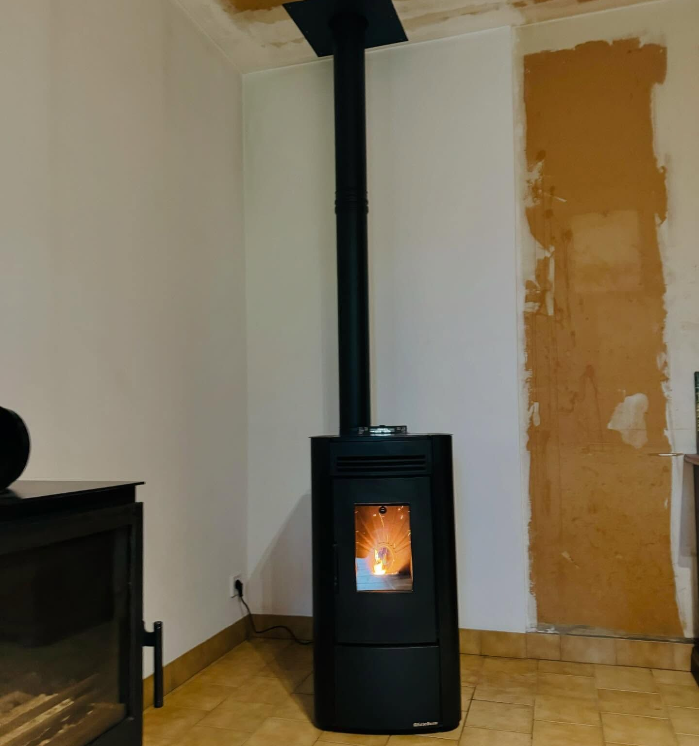 Poêle à granulés noirs avec flammes allumées, conduit de fumée vertical noir et rénovation partielle du mur.