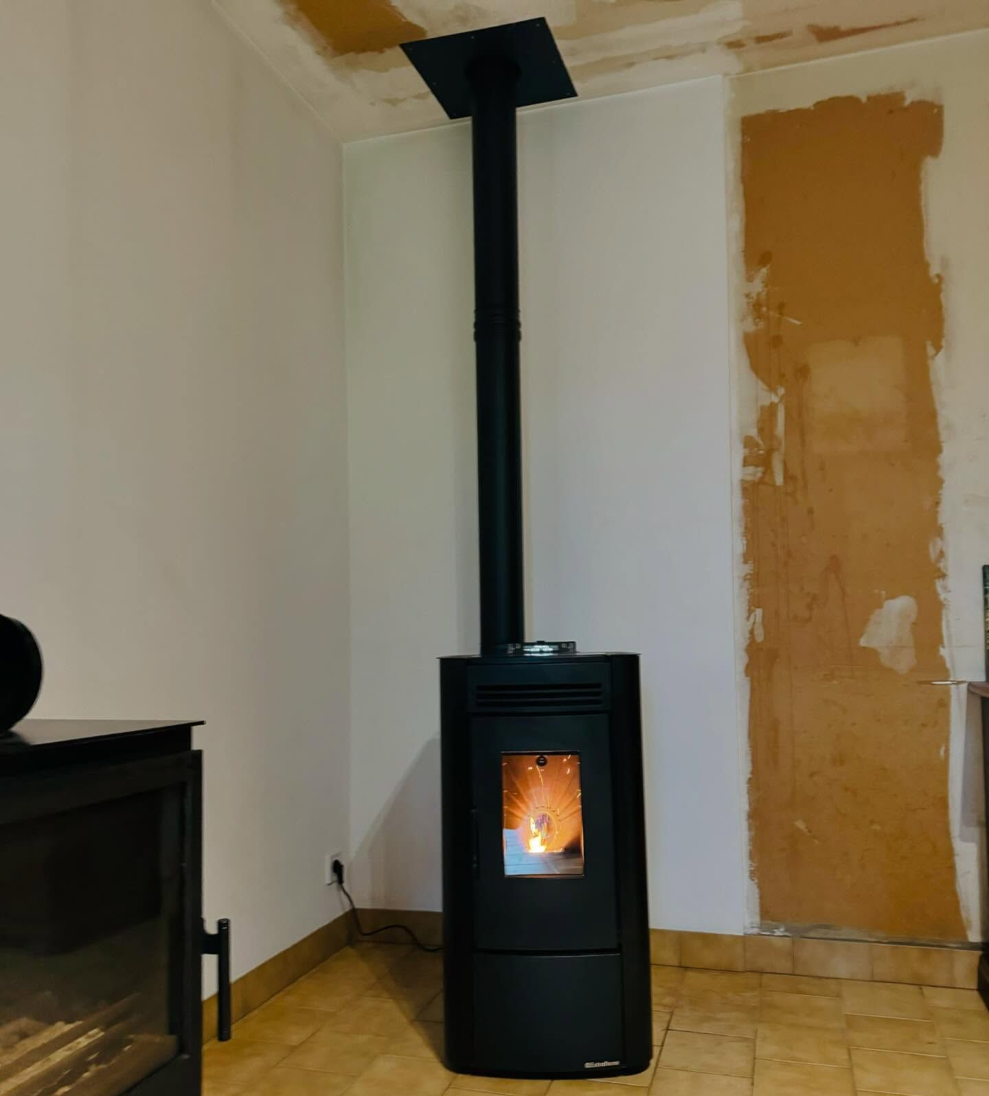 Poêle à granulés noir avec flammes allumées, tuyau noir vertical, parois blanches et partie de mur apparente.