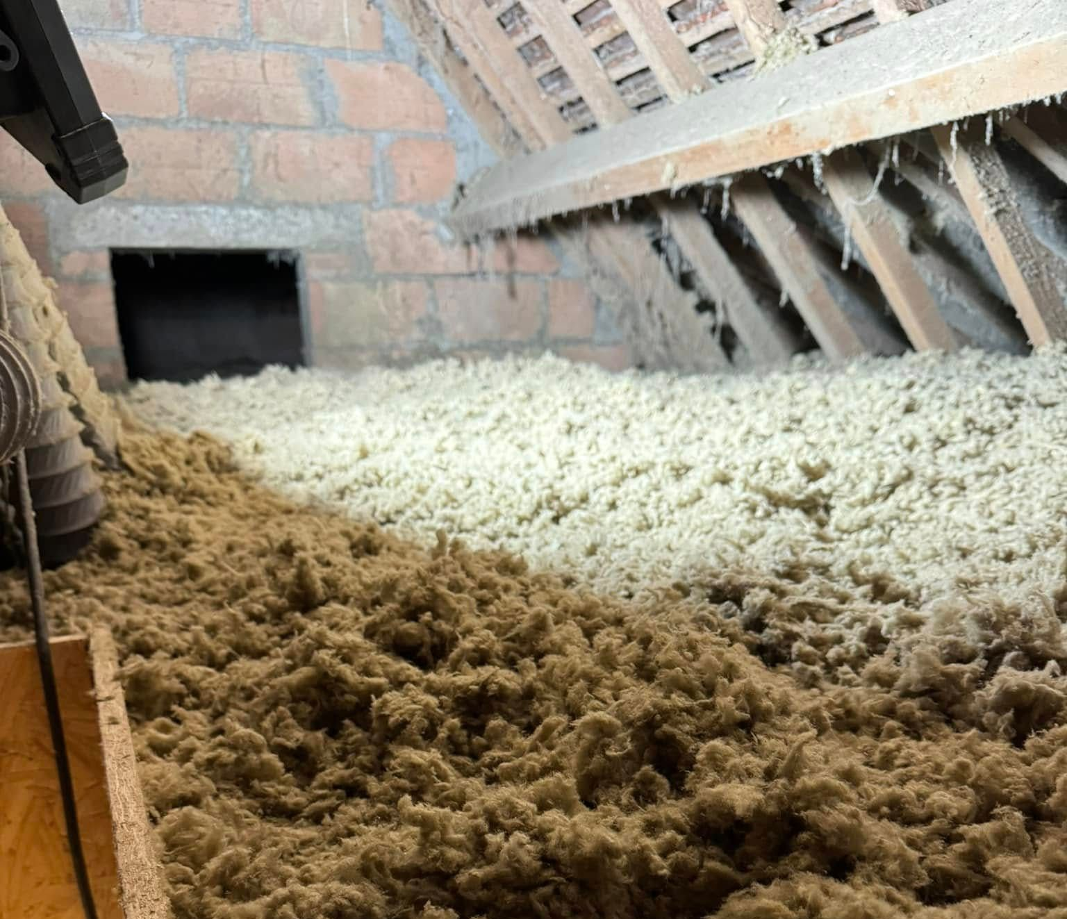 Grenier avec isolation en vrac, mur en briques, poutres en bois et une ouverture sombre.