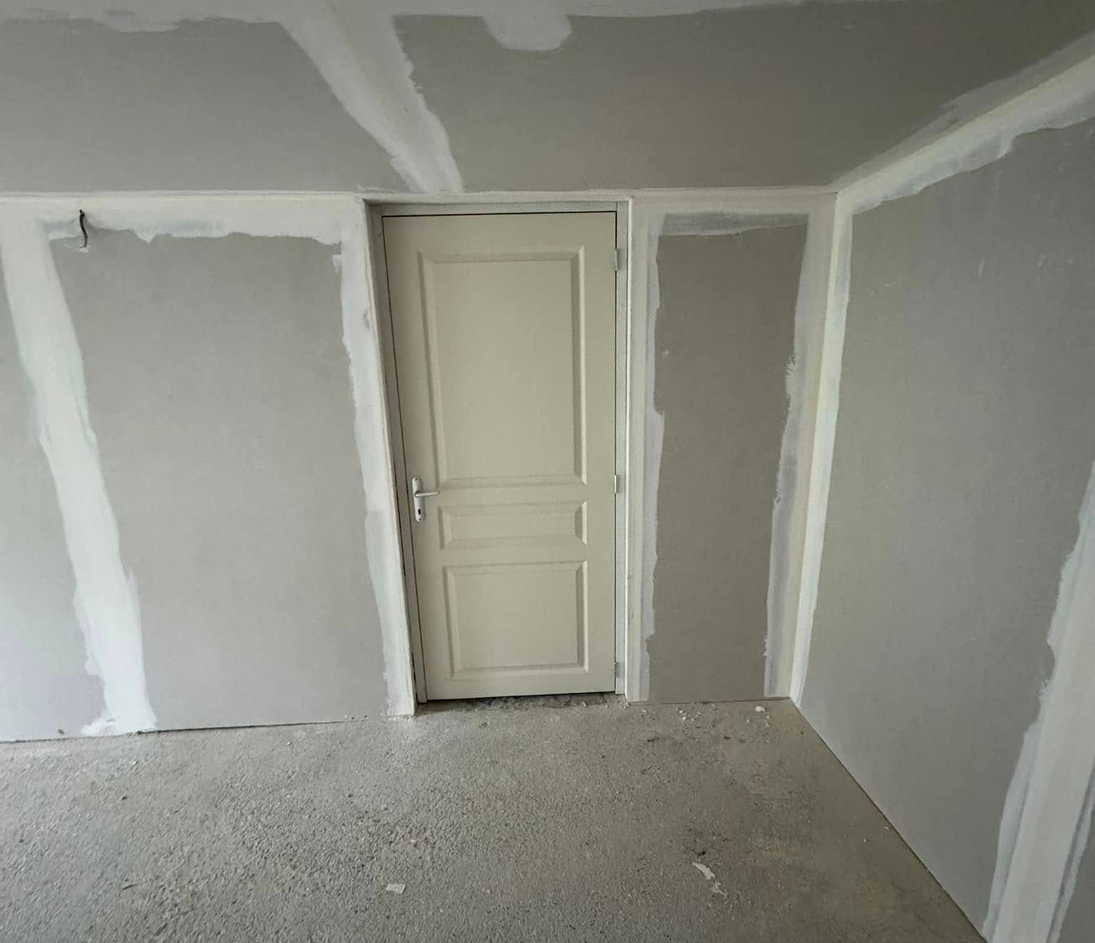 Pièce intérieure avec cloisons sèches non finies autour d'une porte fermée ; murs et sol gris clair.
