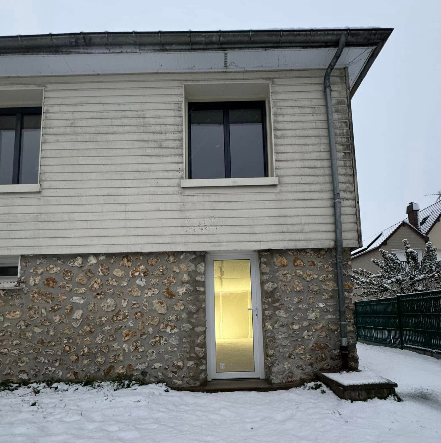 Bâtiment enneigé de deux étages. Bardage blanc au-dessus de fondations en pierre, petite porte illuminée, neige au sol.
