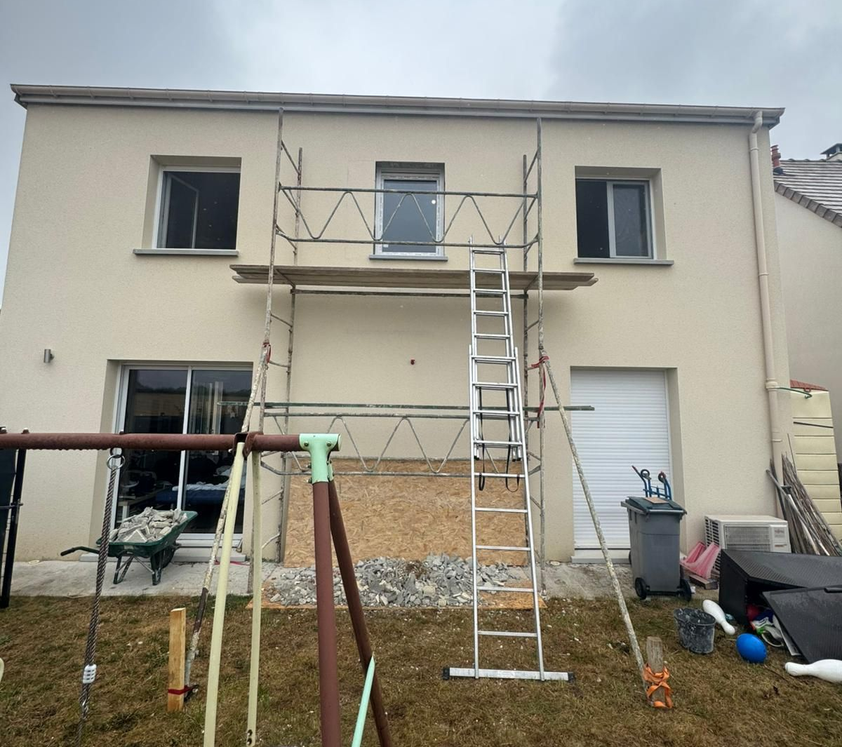Échafaudages sur une maison beige de deux étages en construction, avec une aire de jeux dans la cour.