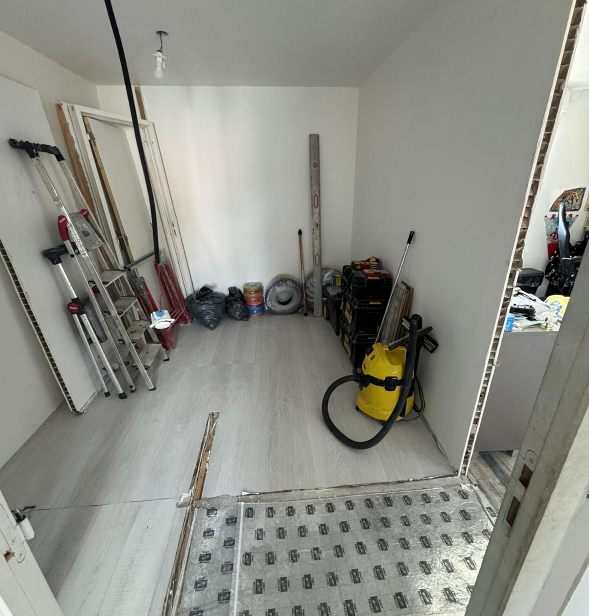 Pièce contenant des matériaux et des outils de construction ; sol brut, murs blancs et éclairage vif.