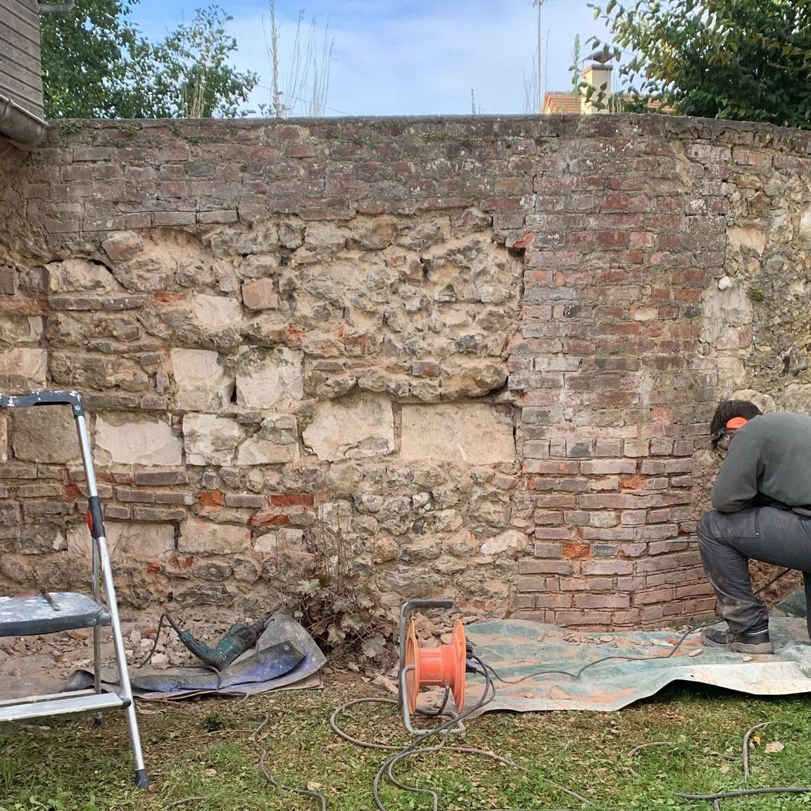 Un homme travaille sur un mur de briques et de pierres érodé par le temps, avec des outils et une échelle au sol.