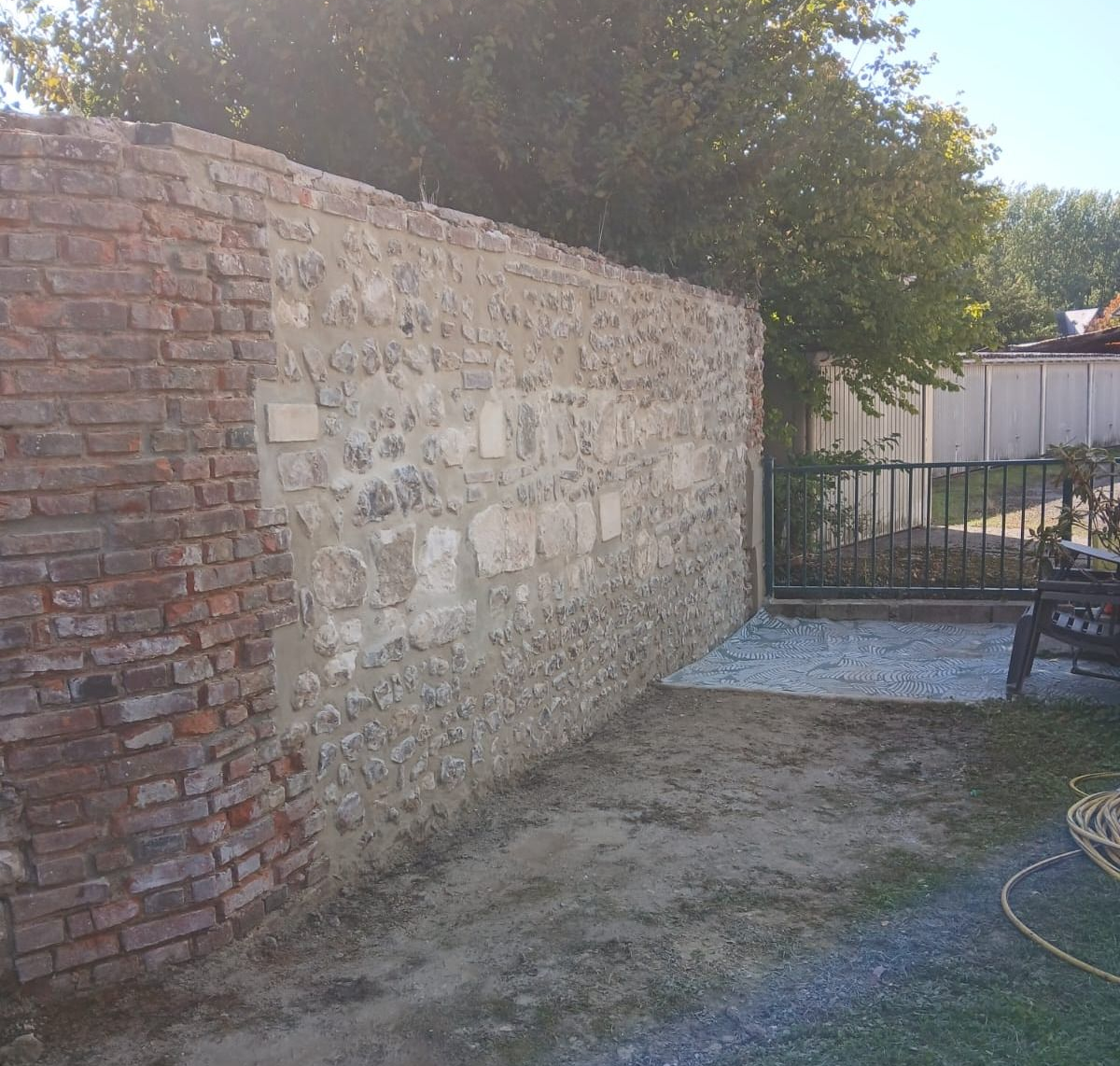 Mur de briques et de pierres dans une cour, avec une zone de gravier et un portail métallique visibles.