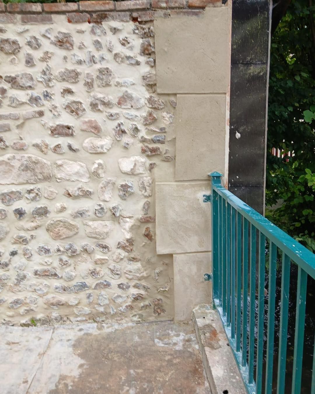 Mur en pierre avec blocs de pierre carrés adjacents, balustrade turquoise et sol en béton.