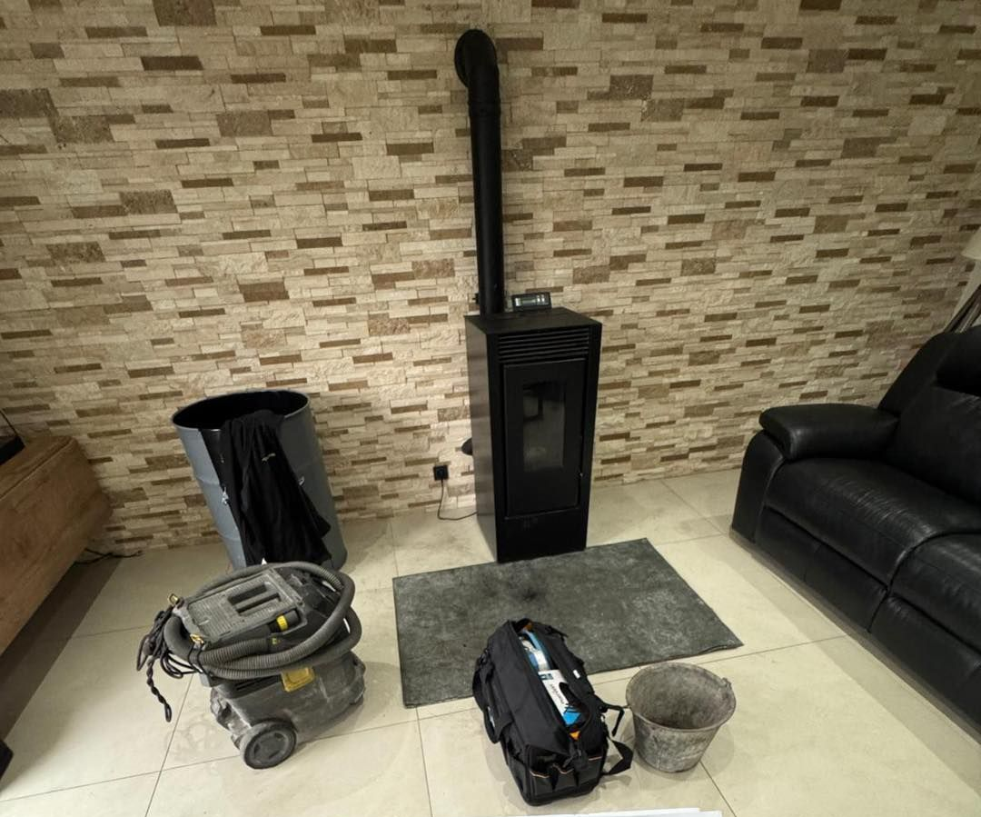 Un poêle noir avec une cheminée se trouve devant un mur en pierre. Un aspirateur, une poubelle et un sac sont à proximité.