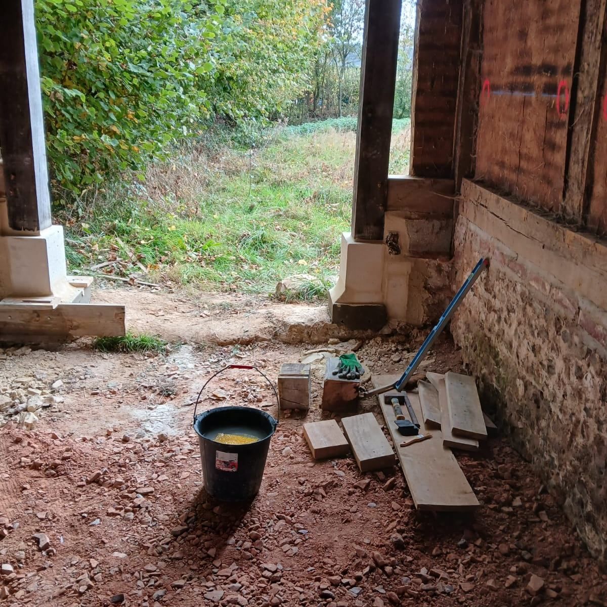 À l'intérieur d'un bâtiment en construction. Des briques, des outils et un seau sont posés sur le sol en terre battue.