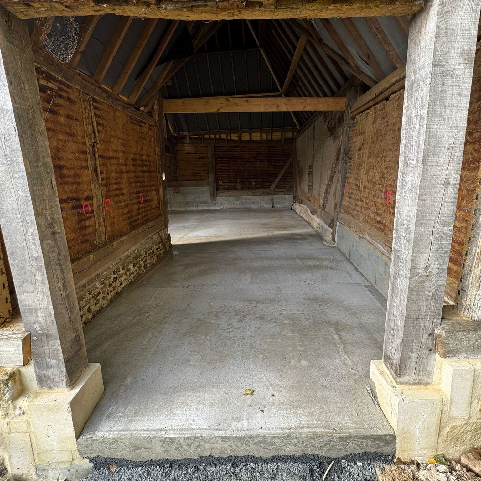 Vue intérieure d'une grange avec un sol en béton fraîchement coulé et des poutres en bois apparentes.