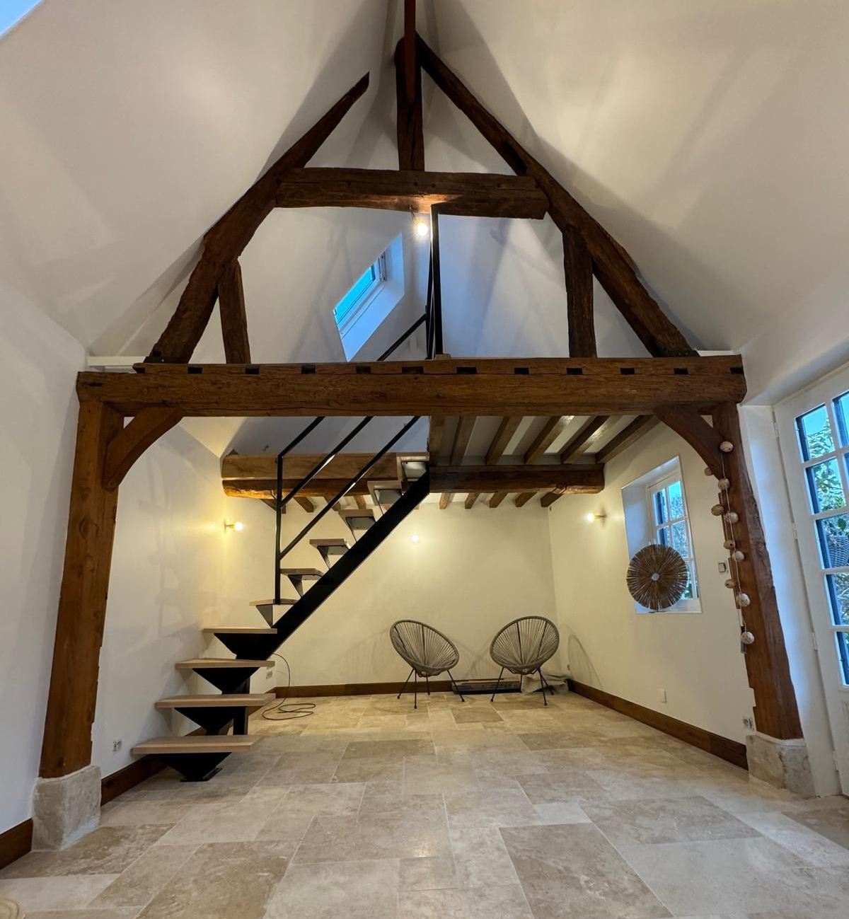 Intérieur avec poutres apparentes, mezzanine, escalier et deux chaises. Murs et sol clairs.