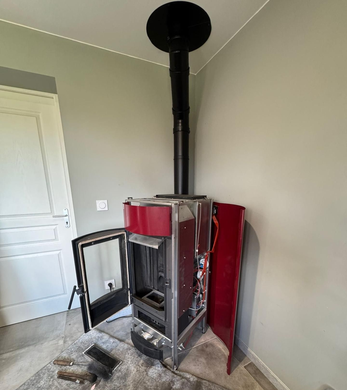 Un poêle à bois avec porte ouverte, panneau rouge, cheminée noire et une brosse dans une pièce à porte blanche.