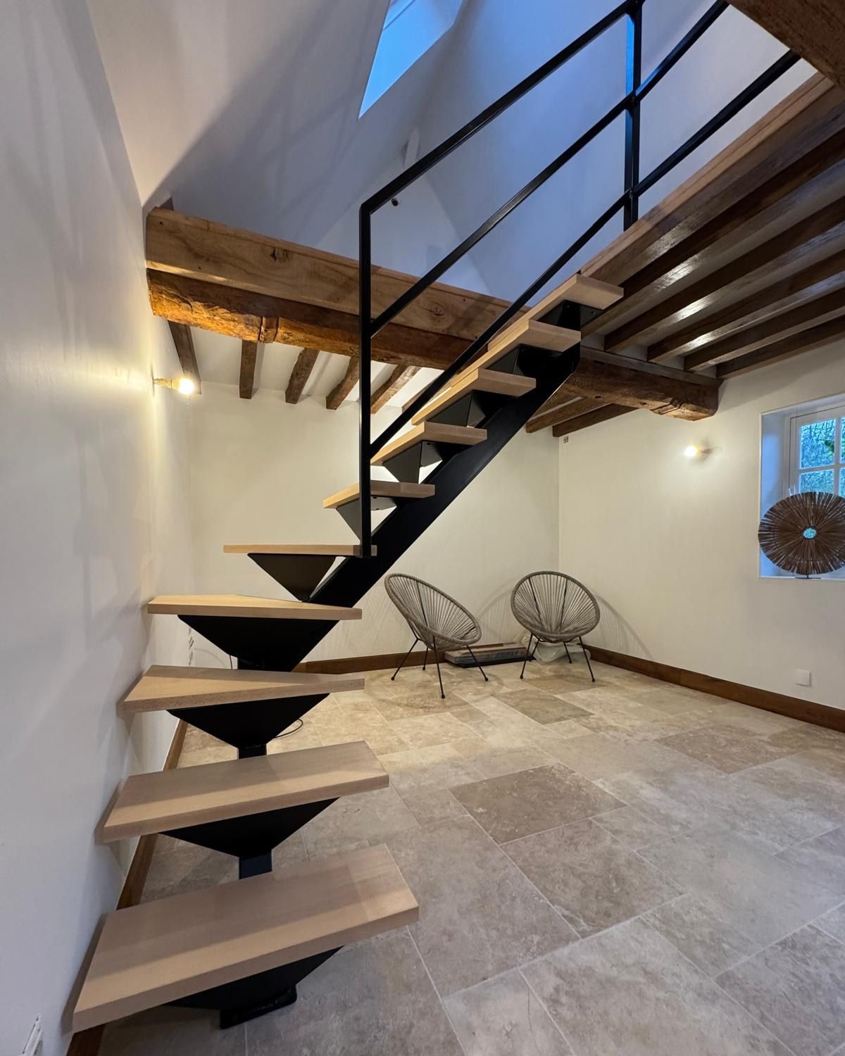 Escalier moderne en bois avec supports en métal noir dans une pièce avec coin salon et poutres apparentes.
