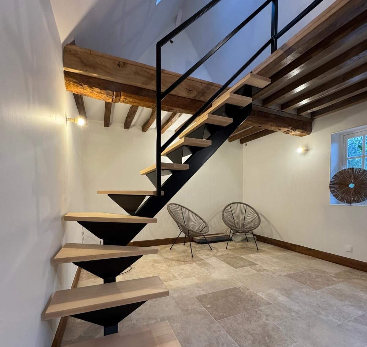 Un escalier aux marches en bois et aux supports noirs mène à une mezzanine. Deux chaises sont placées au pied de l'escalier.