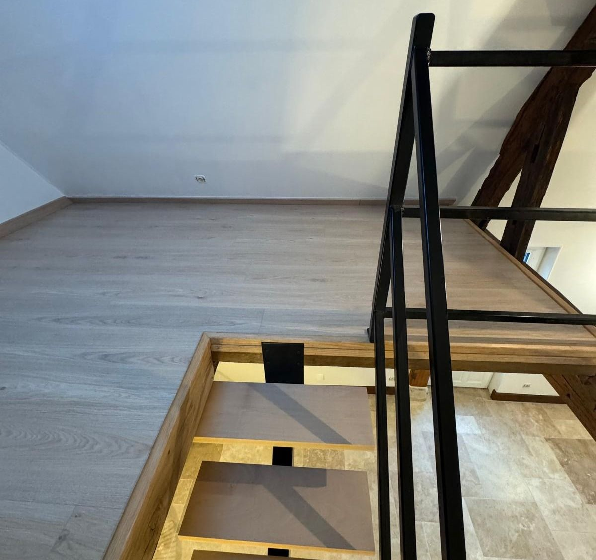 Un escalier en bois avec une rampe noire mène à une mezzanine. On aperçoit des murs beiges et une poutre en bois.