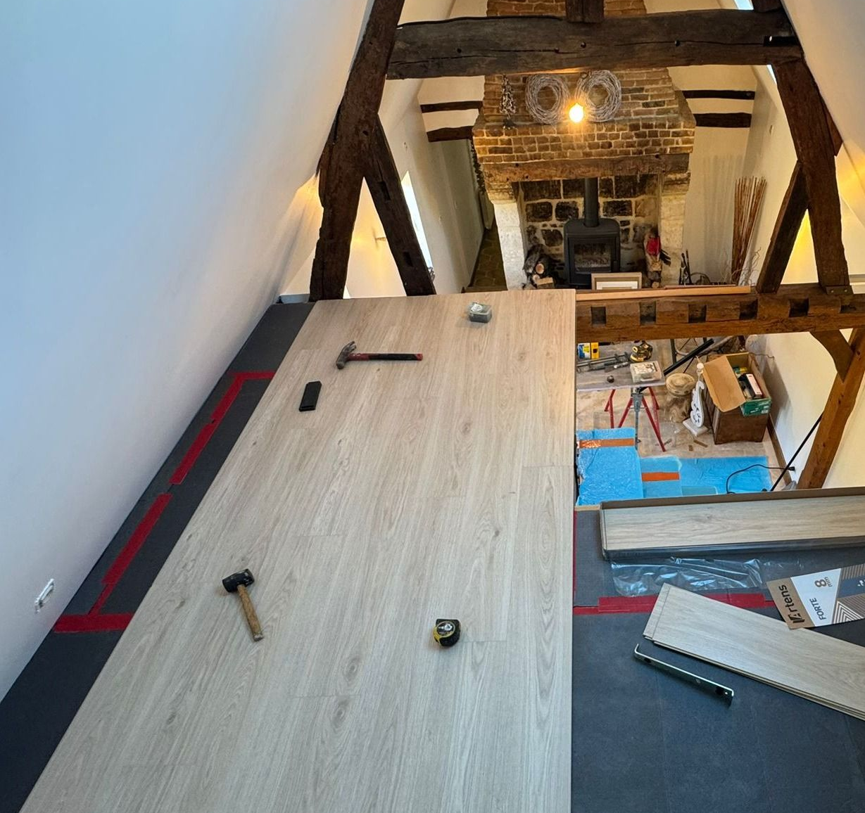 Pose de revêtement de sol dans une pièce avec poutres apparentes et cheminée en briques. Outils et matériaux présents.